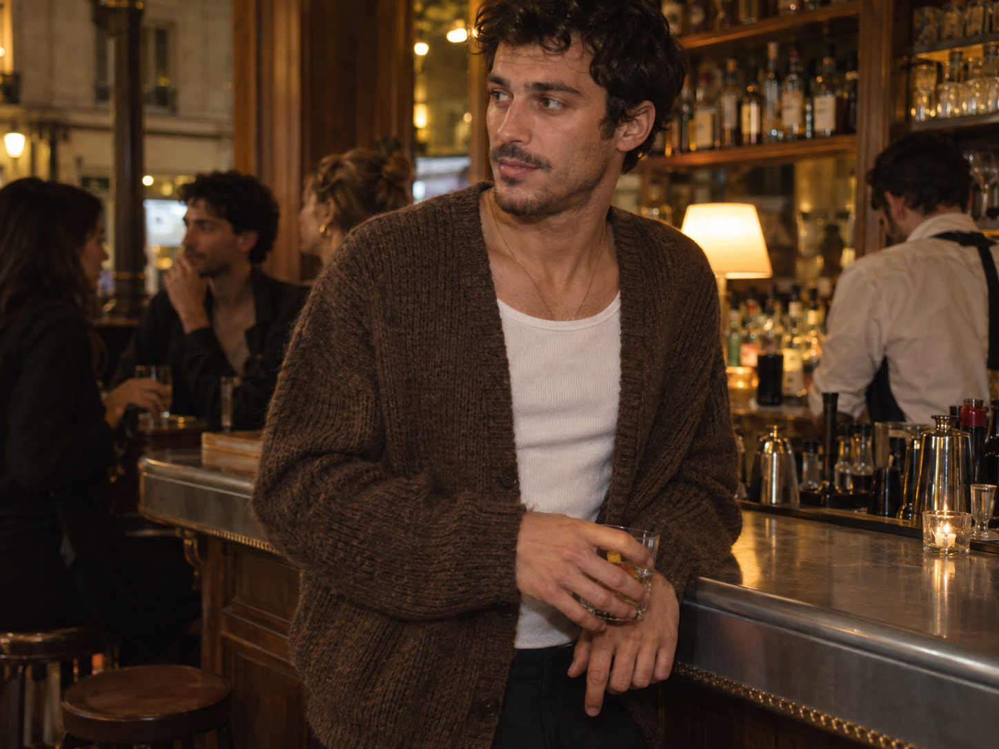 Un jeune homme porte un cardigan en laine sur un débardeur blanc dans un café parisien.