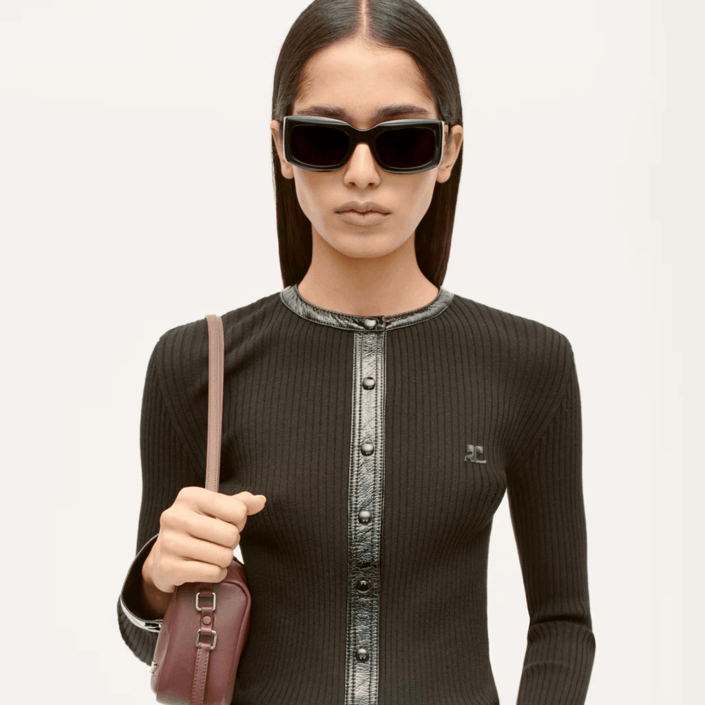 Mannequin portant un cardigan noir Courrèges, des lunettes sombres et un sac bordeaux sur fond clair