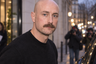 Portrait de Drew Henry en pull noir devant des photographes, dans une rue parisienne à la tombée du jour