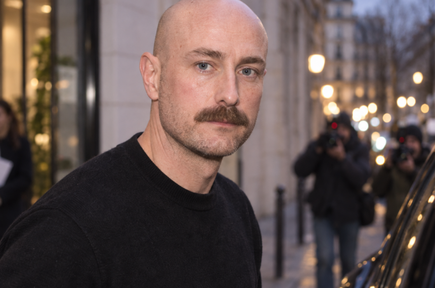 Portrait de Drew Henry en pull noir devant des photographes, dans une rue parisienne à la tombée du jour