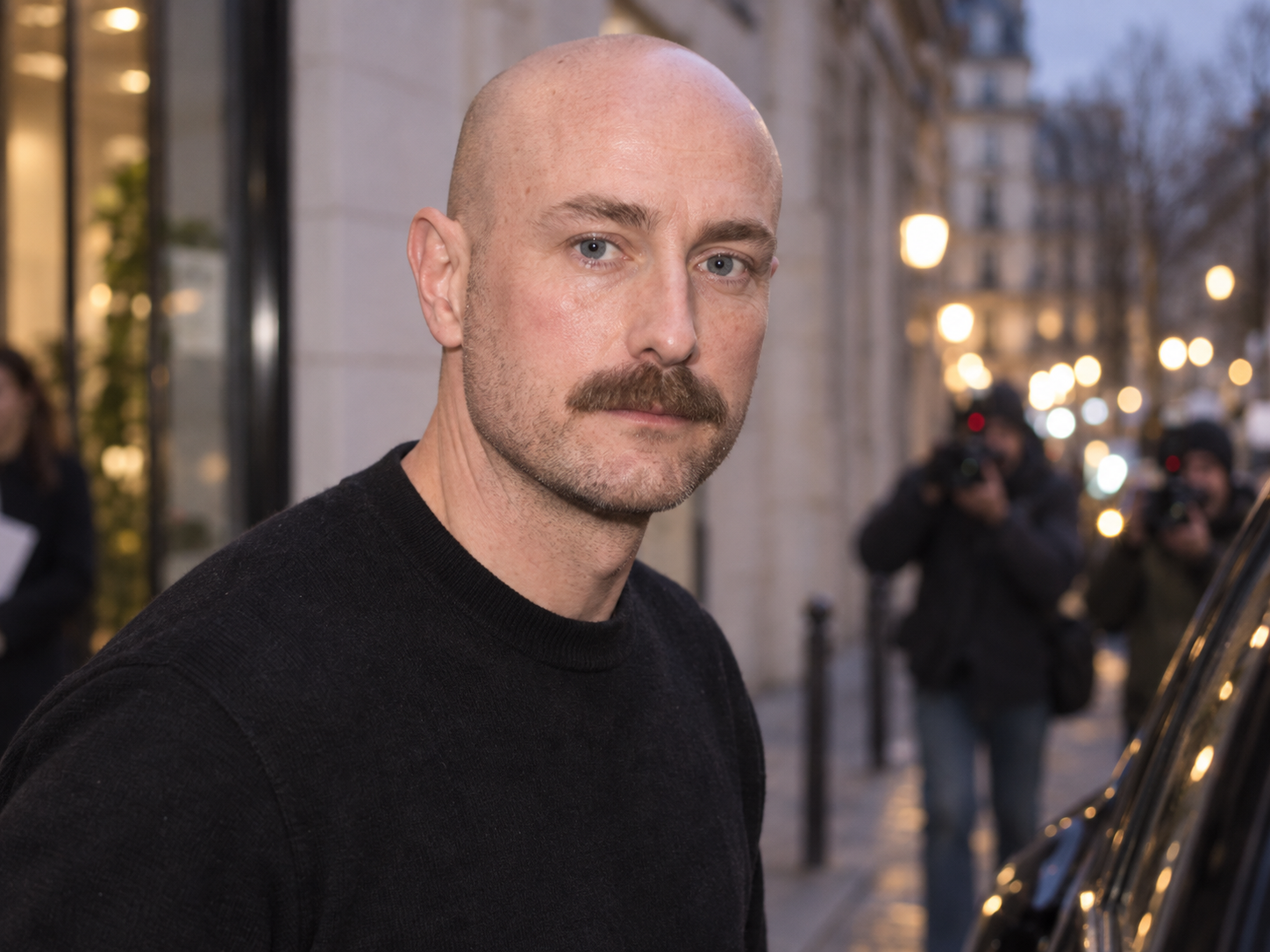 Portrait de Drew Henry en pull noir devant des photographes, dans une rue parisienne à la tombée du jour