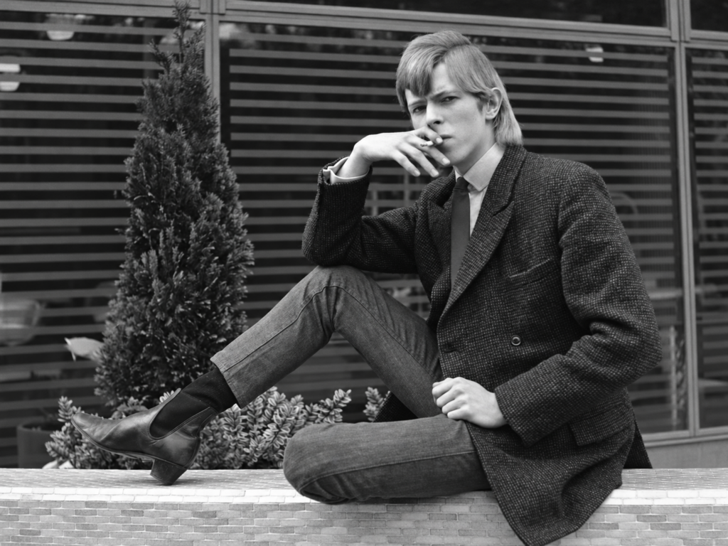 David Bowie assis sur un muret, une cigarette à la main, devant une façade vitrée.