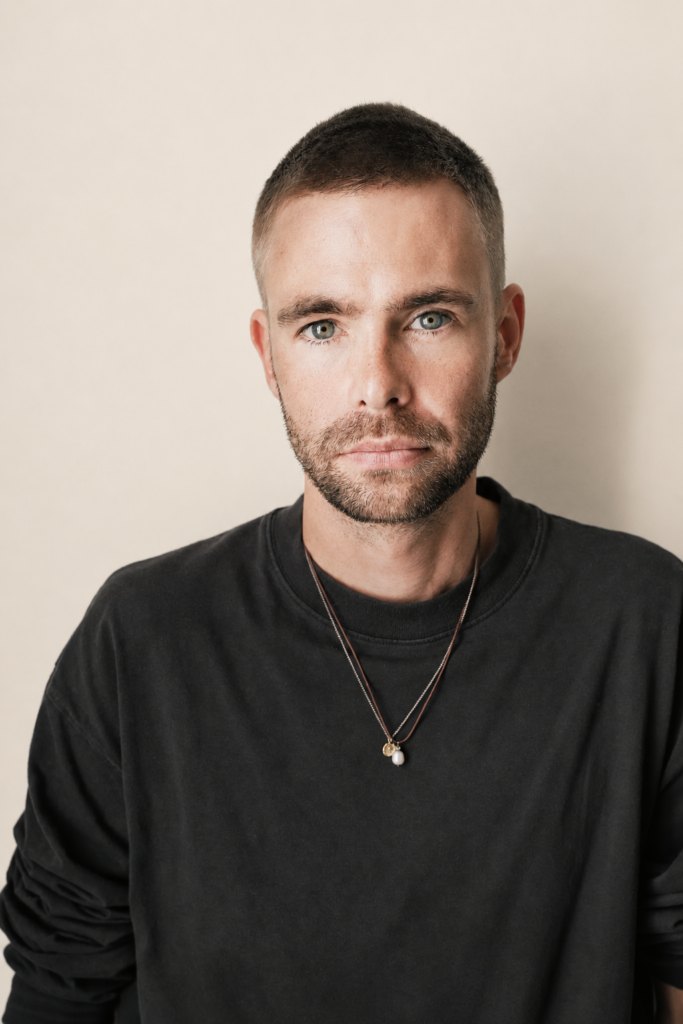 Portrait frontal de Kai Nesselrath en sweat noir, devant un fond beige clair, le regard dirigé vers l’objectif.