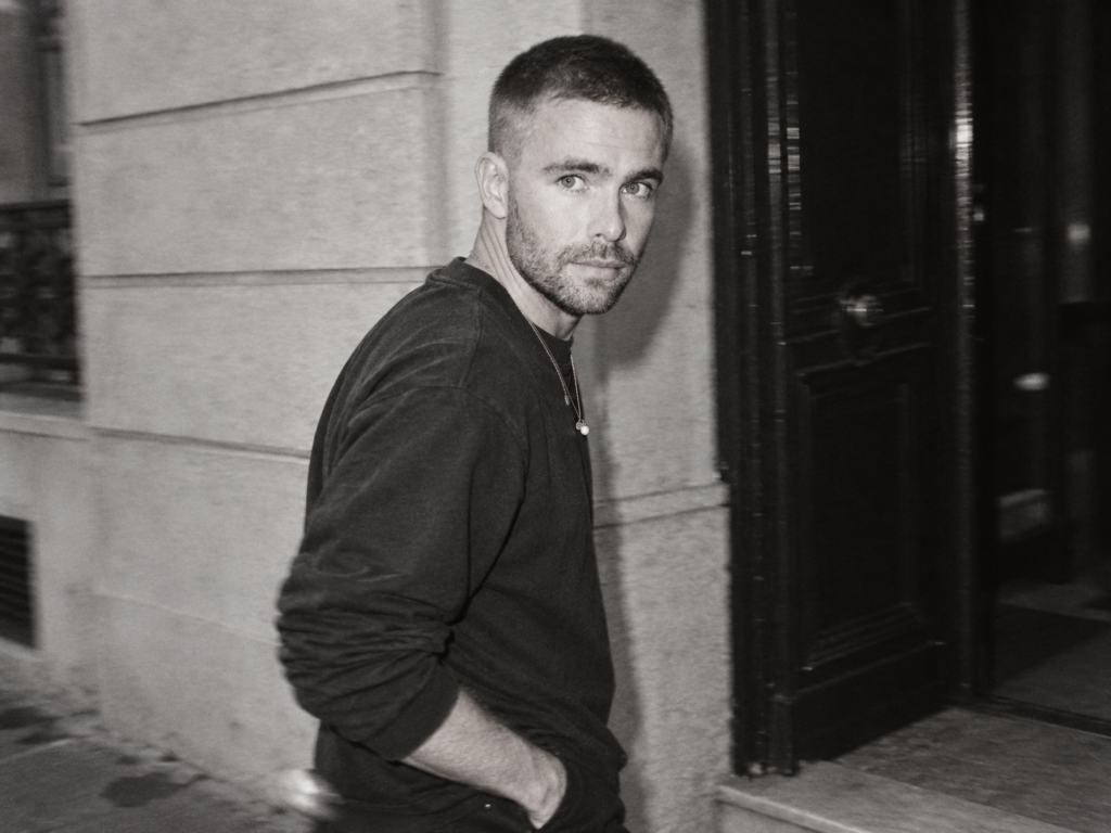 Kai Nesselrath marche devant une façade sombre, vêtu d’un sweat noir, dans un portrait en noir et blanc.