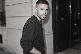 Kai Nesselrath marche devant une façade sombre, vêtu d’un sweat noir, dans un portrait en noir et blanc.