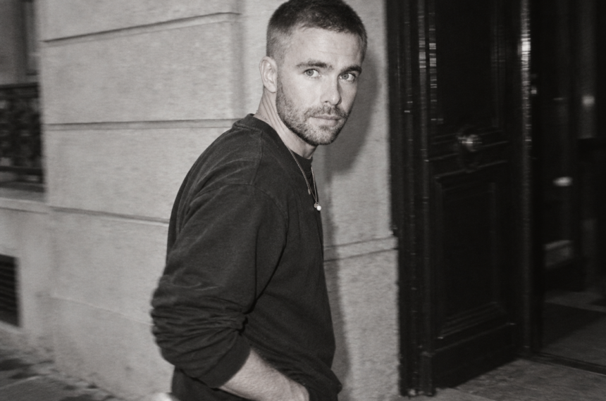 Kai Nesselrath marche devant une façade sombre, vêtu d’un sweat noir, dans un portrait en noir et blanc.
