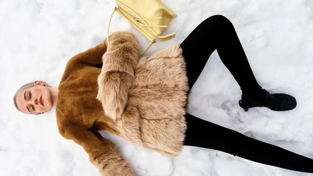 Julia Garner allongée sur la neige, vêtue d’un manteau brun à manches volumineuses, avec un sac jaune Loewe