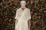 Look Maison Margiela printemps-été 2027 en robe filet écrue et résille sur un podium au décor floral