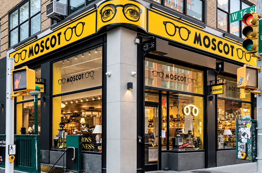 Façade d’une boutique Moscot à un angle de rue, avec son enseigne jaune et ses lunettes visibles derrière les vitrines.