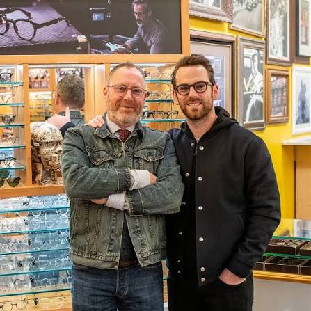 Harvey et Zack Moscot posent ensemble dans la boutique de SoHo, devant des présentoirs de lunettes et des portraits encadrés.