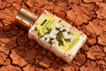 Flacon du parfum Tears couché sur une terre rouge craquelée, avec des traces de poussière sur le bouchon et l’étiquette.