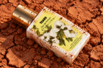 Flacon du parfum Tears couché sur une terre rouge craquelée, avec des traces de poussière sur le bouchon et l’étiquette.