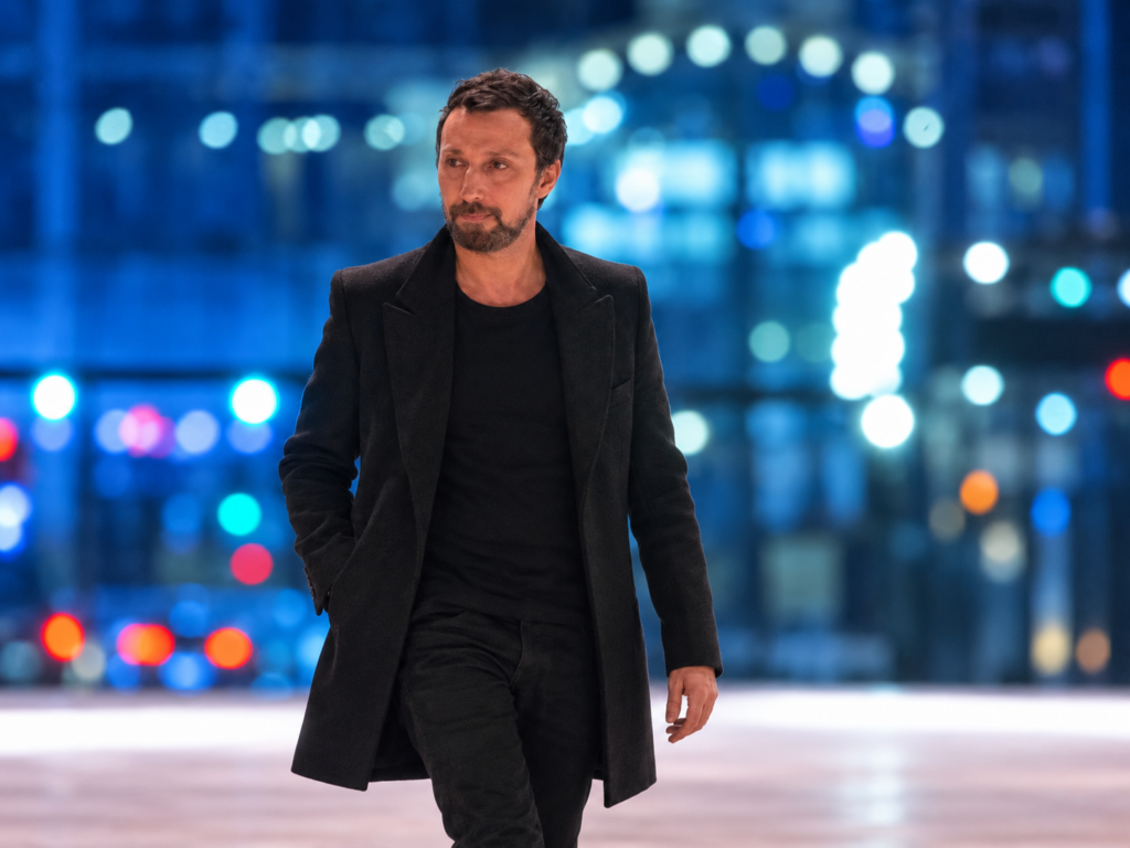 Anthony Vaccarello marche sur le podium Saint Laurent, vêtu de noir, devant des lumières bleues floues.