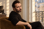 Portrait d’Anthony Vaccarello assis près d’une fenêtre, vêtu de noir.