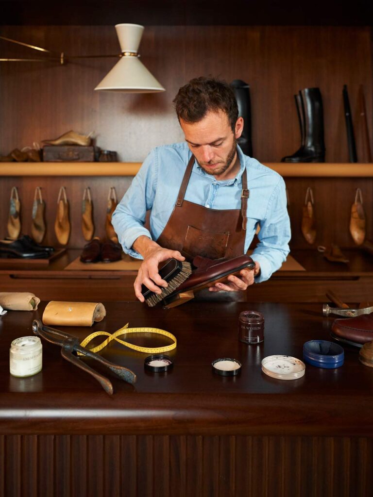 Un homme brosse un soulier en cuir marron J.M. Weston derrière un comptoir, entouré d’outils et de produits d’entretien.