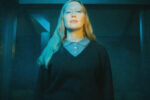 Julia Jacklin face à l’objectif, les cheveux longs et un haut sombre, sous une lumière bleue et jaune.