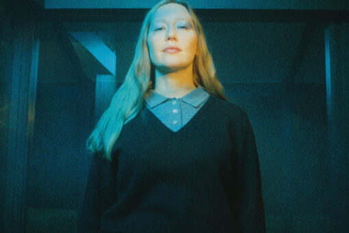 Julia Jacklin face à l’objectif, les cheveux longs et un haut sombre, sous une lumière bleue et jaune.
