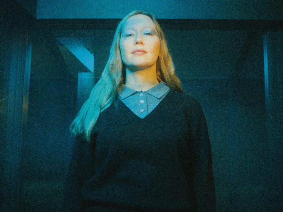 Julia Jacklin face à l’objectif, les cheveux longs et un haut sombre, sous une lumière bleue et jaune.