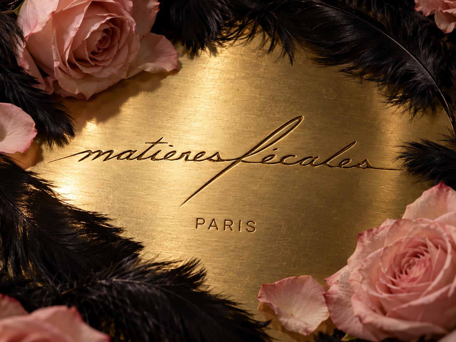 Logo Matières Fécales gravé sur une surface dorée, entouré de plumes noires et de roses rose pâle.
