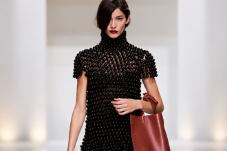 Mannequin du défilé Celine à Paris en robe noire texturée, collants noirs et grand sac bordeaux.