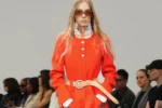 Silhouette Courrèges en manteau orange ceinturé, col montant blanc et lunettes rouges lors du défilé à Paris