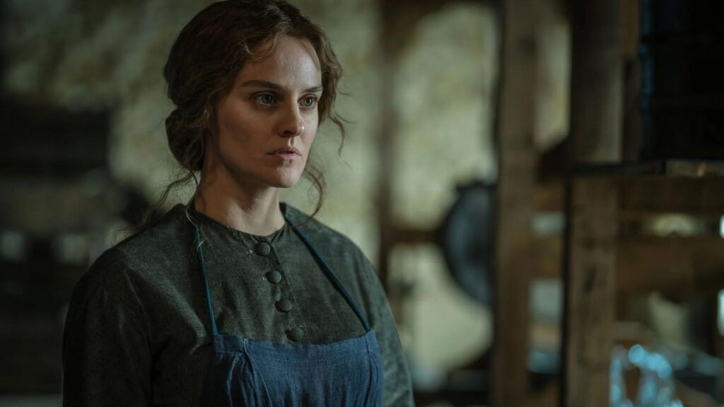Fantine, jouée par Noémie Merlant dans Les Misérables, porte une robe grise boutonnée et un tablier bleu.