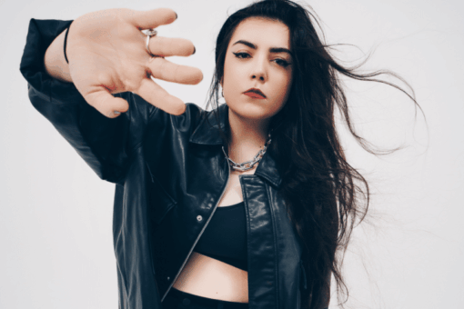Hoshi, chanteuse française, en veste de cuir noir, cheveux au vent et main tendue vers l’objectif sur fond blanc.