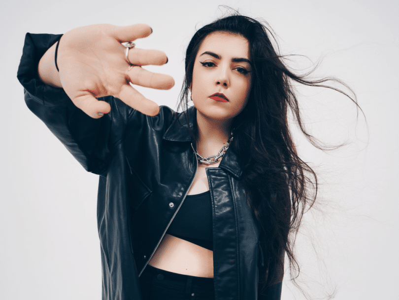 Hoshi, chanteuse française, en veste de cuir noir, cheveux au vent et main tendue vers l’objectif sur fond blanc.