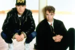 Chris Lowe et Neil Tennant des Pet Shop Boys, vêtus de noir, avec une casquette BOY, dans un intérieur au parquet en bois.
