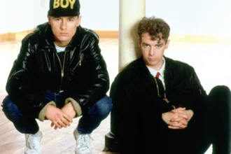 Chris Lowe et Neil Tennant des Pet Shop Boys, vêtus de noir, avec une casquette BOY, dans un intérieur au parquet en bois.