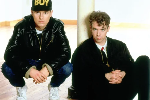 Chris Lowe et Neil Tennant des Pet Shop Boys, vêtus de noir, avec une casquette BOY, dans un intérieur au parquet en bois.
