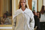Mannequin du défilé The Row printemps-été 2027 en manteau gris clair, col en dentelle blanche et pantalon noir.