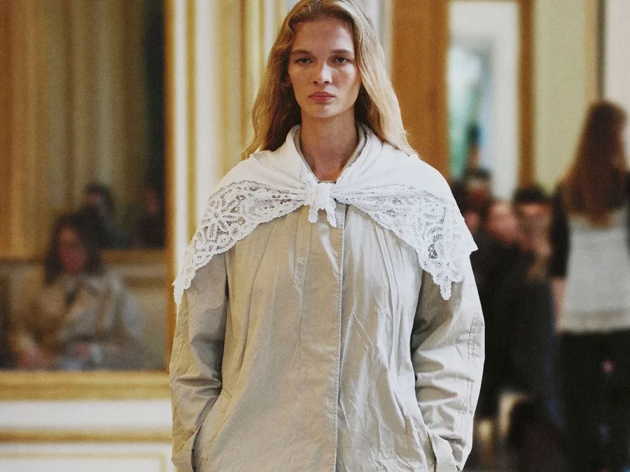 Mannequin du défilé The Row printemps-été 2027 en manteau gris clair, col en dentelle blanche et pantalon noir.