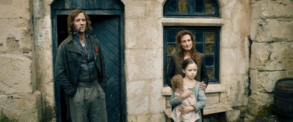 Les Thénardier dans Les Misérables portent des vêtements sombres devant une maison de pierre, avec une fillette et sa poupée.