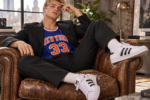 Jeune homme blond portant des Adidas Superstar blanches et noires, un costume noir et un maillot de basket New York 33, assis dans un fauteuil en cuir vintage.