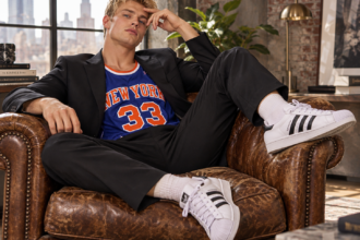 Jeune homme blond portant des Adidas Superstar blanches et noires, un costume noir et un maillot de basket New York 33, assis dans un fauteuil en cuir vintage.