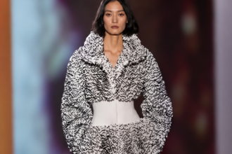 Mannequin du défilé Alaïa printemps-été 2027 à Paris portant un long manteau noir et blanc texturé, cintré par une large ceinture blanche.