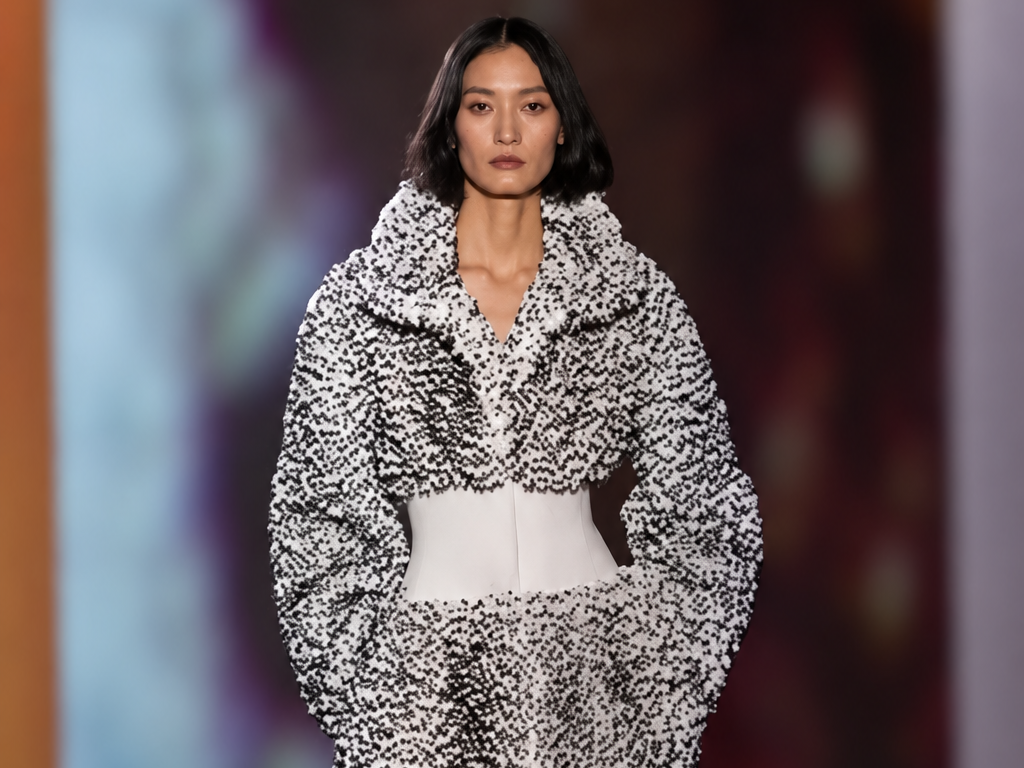 Mannequin du défilé Alaïa printemps-été 2027 à Paris portant un long manteau noir et blanc texturé, cintré par une large ceinture blanche.