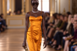 Silhouette du défilé Balmain printemps-été 2027 à la Paris Fashion Week, dans une robe satinée orange portée avec des lunettes noires.