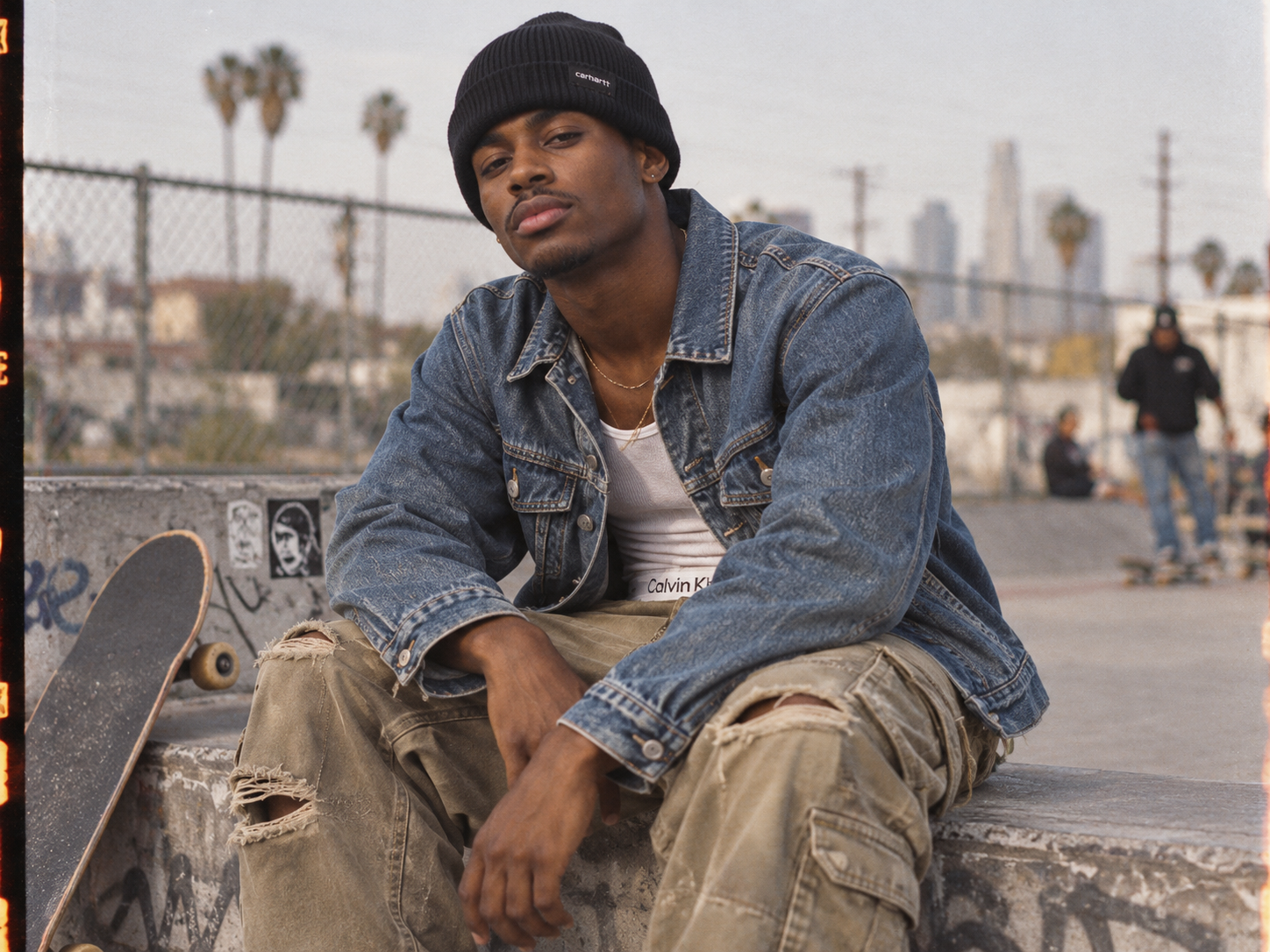 Jeune homme noir portant un bonnet Carhartt WIP noir, une veste en jean et un pantalon cargo kaki déchiré dans un skatepark urbain.