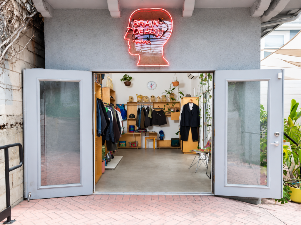 Entrée d'une boutique Brain Dead avec son enseigne lumineuse rouge en forme de tête, vêtements et objets exposés à l'intérieur.