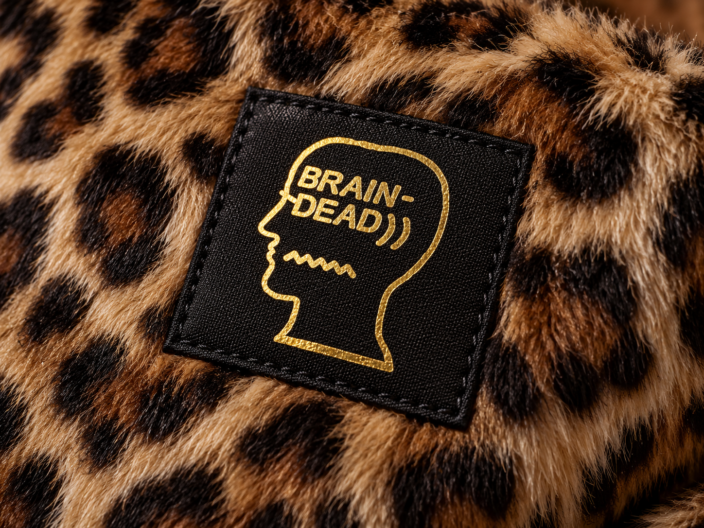 Logo Brain Dead imprimé en doré sur une étiquette noire cousue sur un tissu en fausse fourrure léopard.
