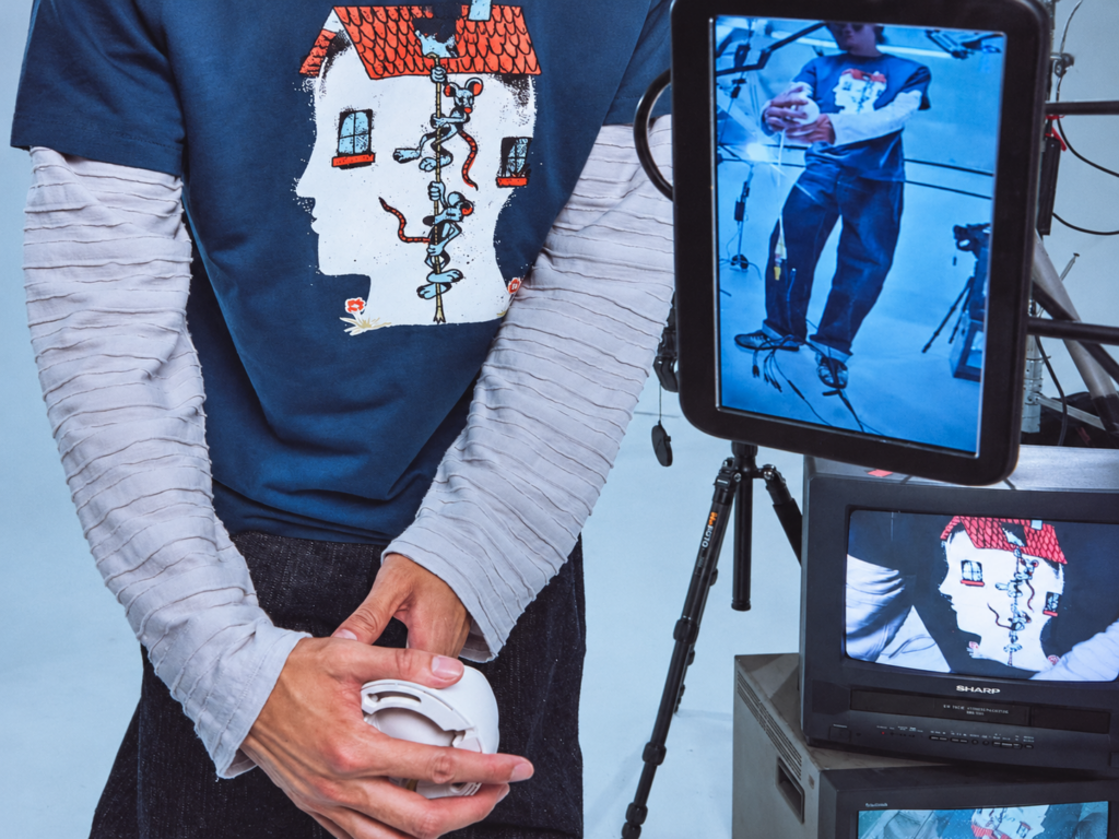 Tee-shirt bleu Brain Dead à imprimé graphique porté sur un haut à manches longues, photographié devant plusieurs écrans vidéo.