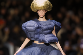 Mannequin du défilé Comme des Garçons printemps-été 2027 portant un ensemble bleu marine aux volumes matelassés et un imposant chapeau doré.