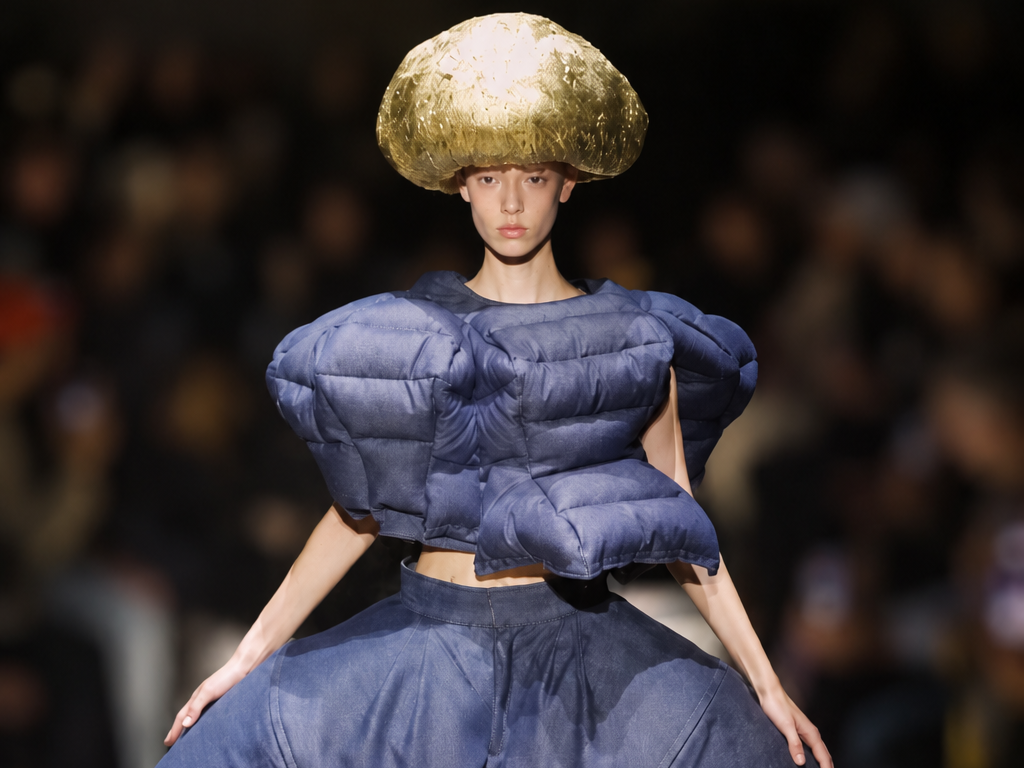Mannequin du défilé Comme des Garçons printemps-été 2027 portant un ensemble bleu marine aux volumes matelassés et un imposant chapeau doré.