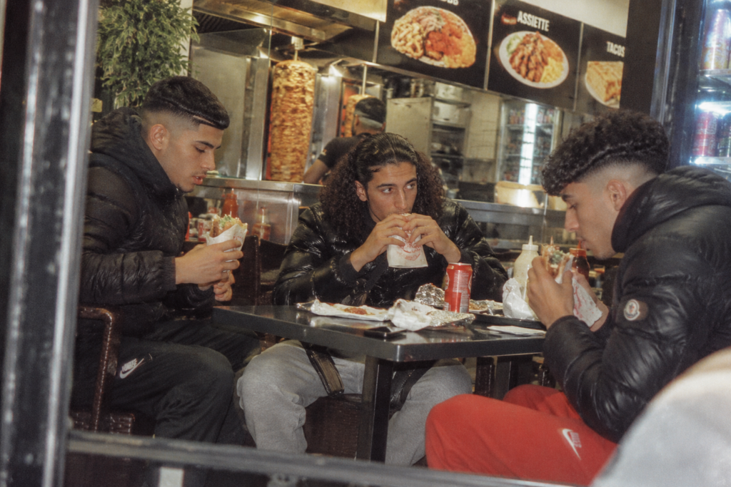 Trois jeunes hommes en doudounes noires et joggings gris, rouge et noir mangent des kebabs dans un restaurant.