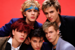 Duran Duran dans les années 1980, les cinq musiciens réunis devant un fond rouge, avec leurs coiffures et tenues new wave.