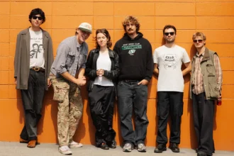 Six musiciens du groupe britannique Fat Dog devant un mur orange, vêtus de tenues décontractées et de lunettes de soleil.