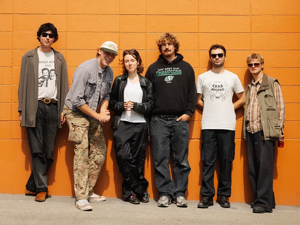 Six musiciens du groupe britannique Fat Dog devant un mur orange, vêtus de tenues décontractées et de lunettes de soleil.