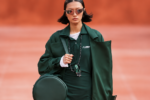 Silhouette verte du défilé Lacoste printemps-été 2027 sur podium terracotta, avec manteau long, mini-robe, lunettes de soleil et sac rond.